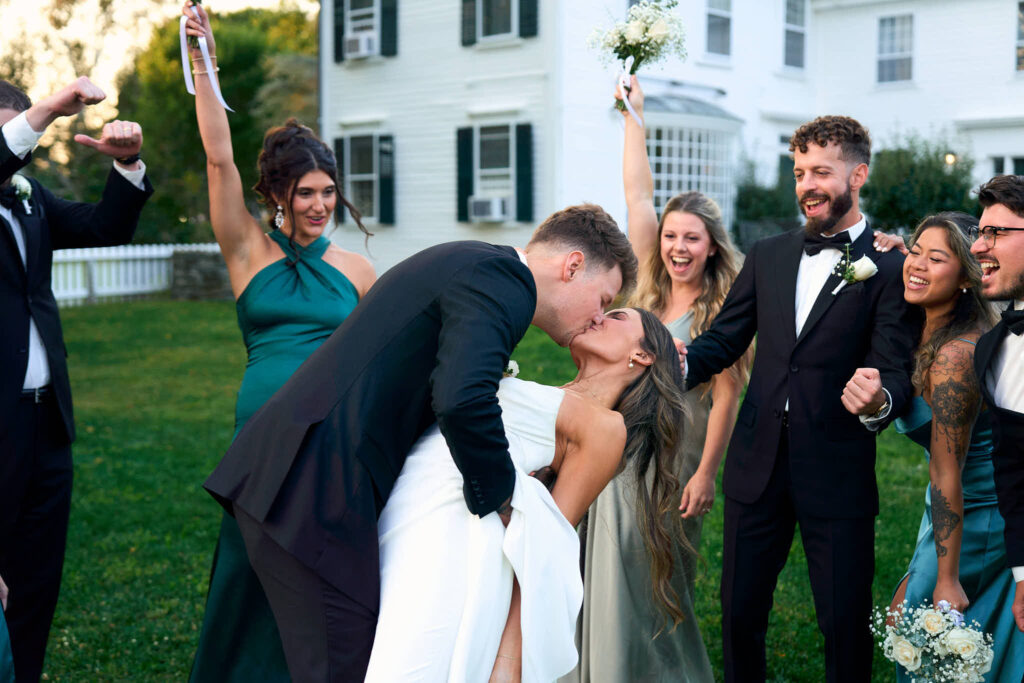 Bride and groom kiss with bridal party cheering them on in front of Mount Hope Farm.